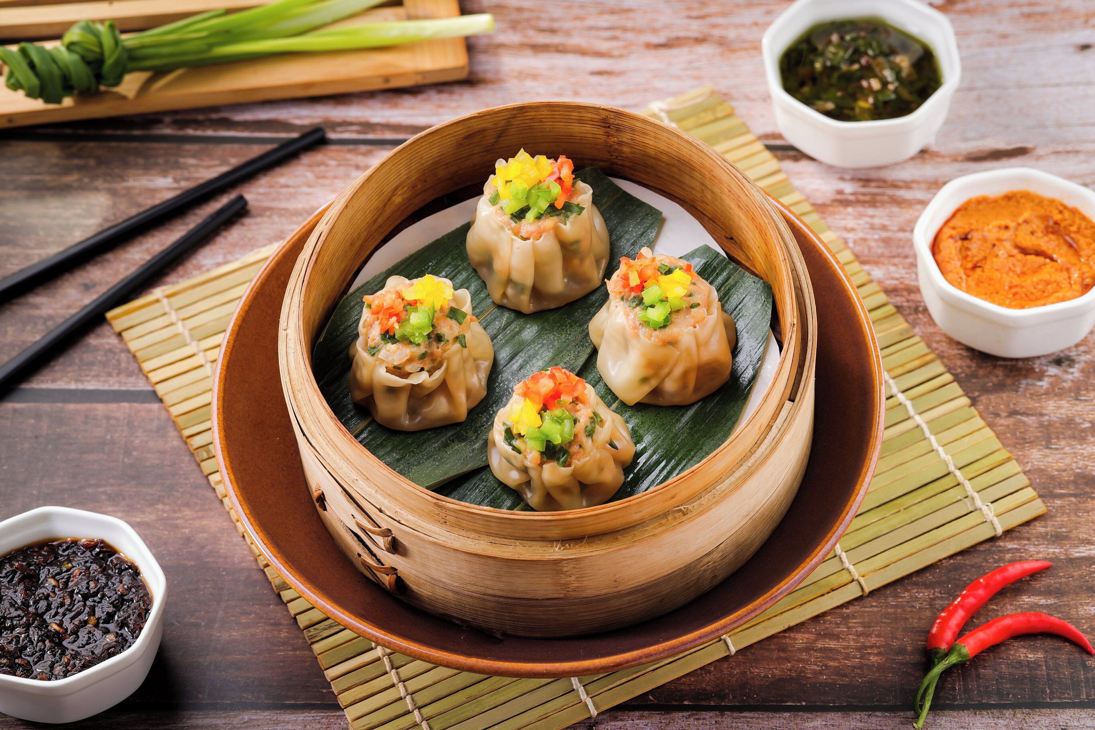 Uma cesta de bambu com dumplings sobre uma mesa de madeira, acompanhada por pequenas tigelas de molhos para acompanhar e pauzinhos.