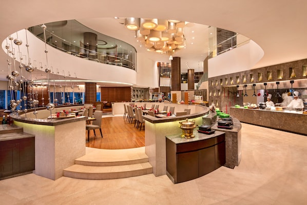 Un hall d’hôtel moderne doté d’un bar central, de différents espaces aménagés pour s’asseoir et d’un plafond haut orné de luminaires originaux.