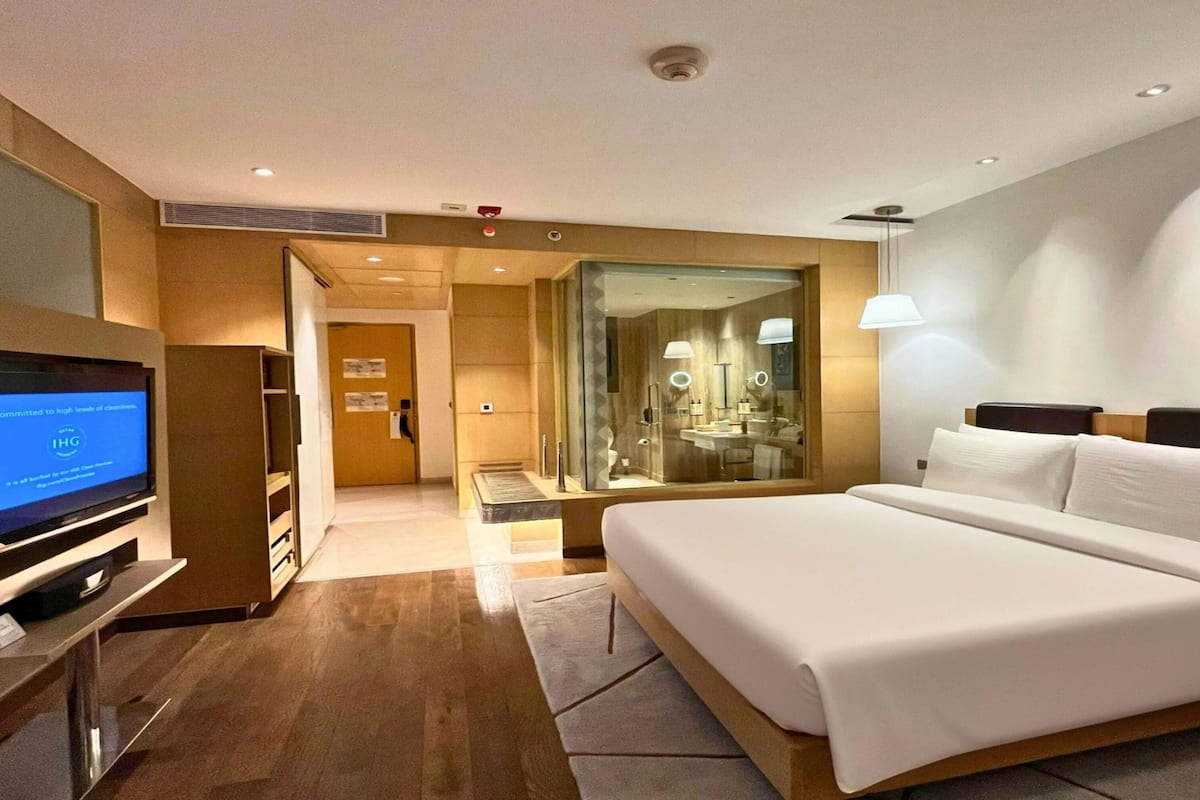Una habitación de hotel moderna con una cama grande, televisión, baño con paredes de vidrio y piso de madera.