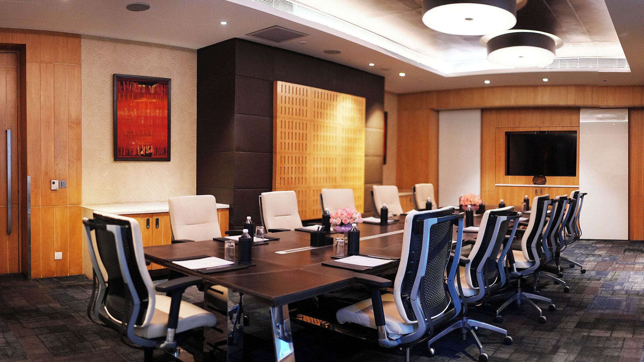 A conference room with a long table, multiple chairs, a television screen, and a decorative panel on the wall.