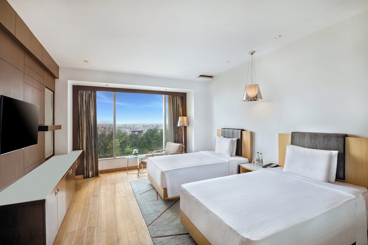 A hotel room with two beds, a TV, a desk, and a large window with a city view.