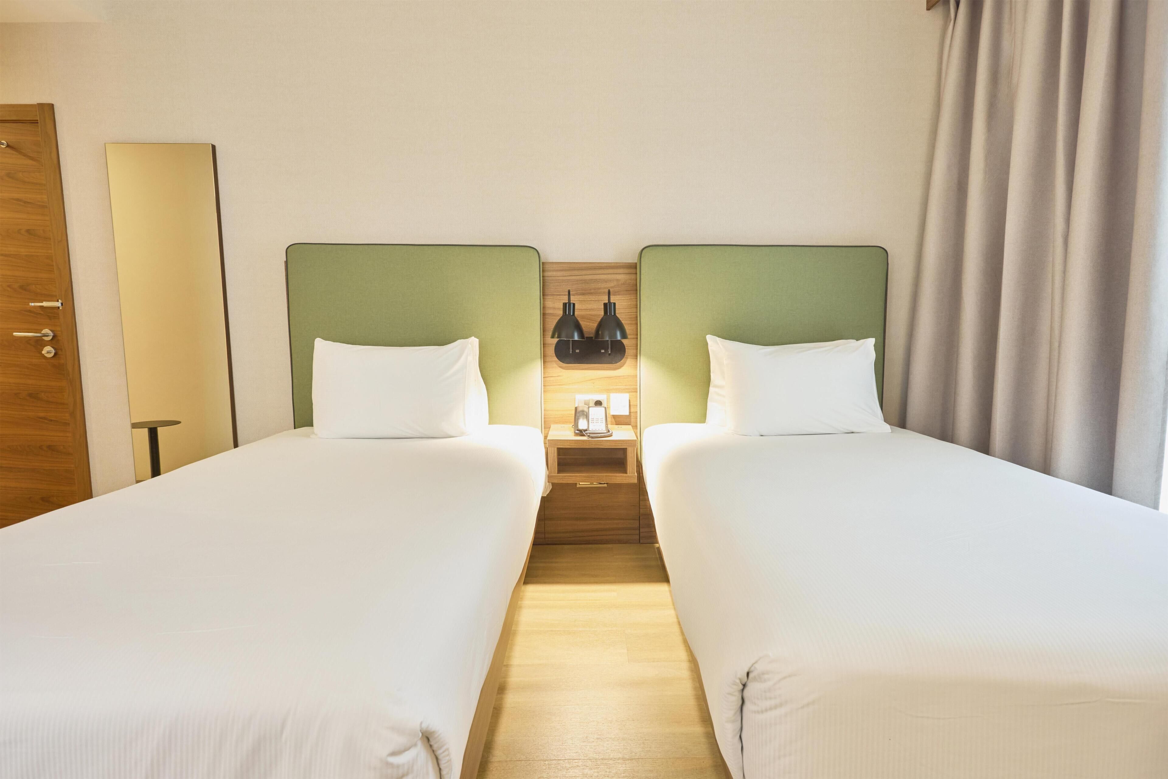 Una habitación de hotel con dos camas individuales, una mesita de noche de madera con una lámpara y una puerta de madera.