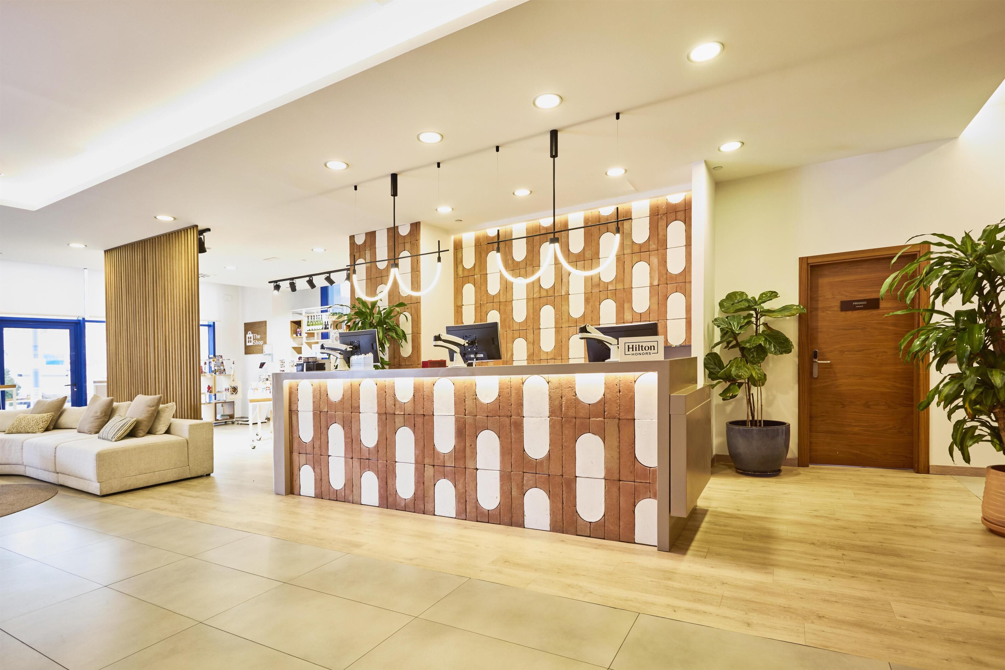 Zona de recepción de hotel con diseño moderno, un mostrador de recepción, un área de estar con sofá y plantas en macetas.