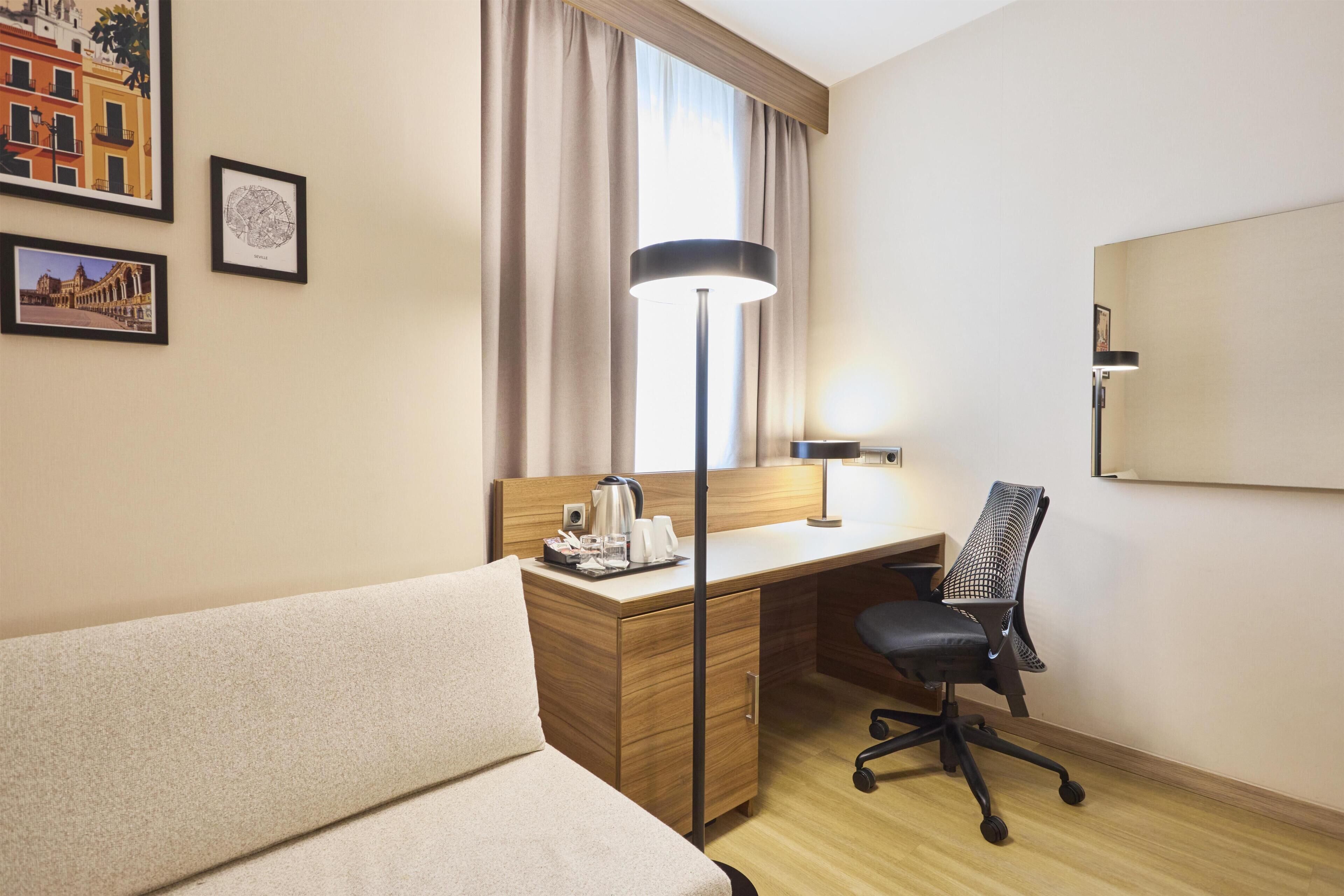 A hotel room with a bed, desk, chair, lamp, and framed pictures on the wall.
