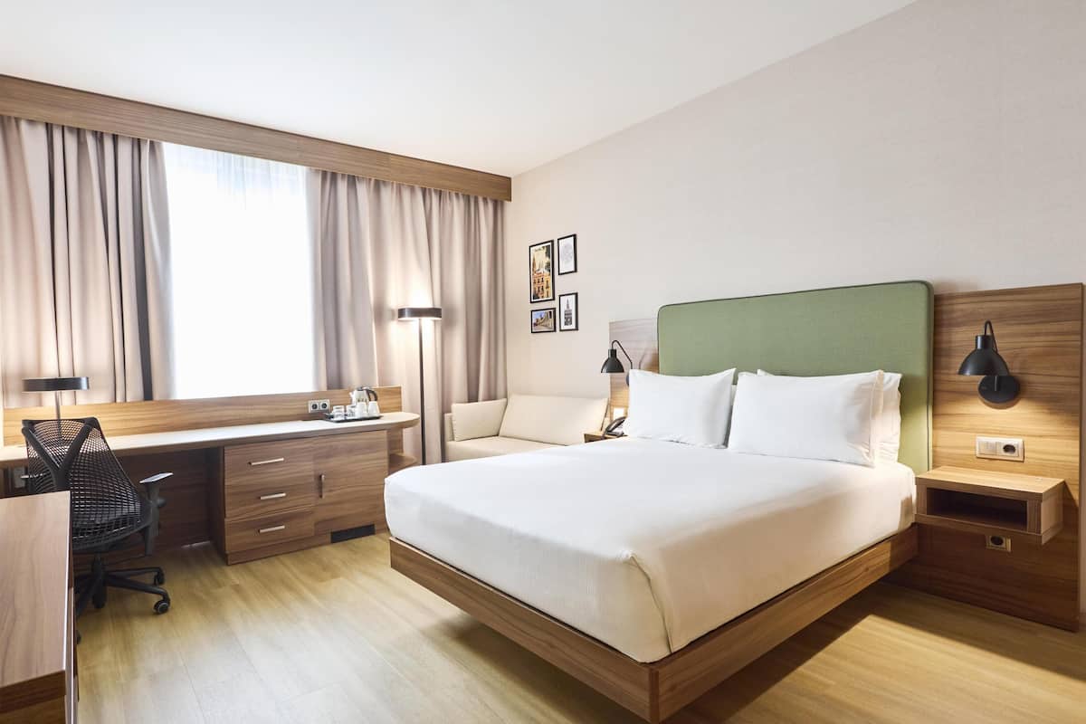 A hotel room with a bed, desk, chair, bedside tables, lamp, and curtains.