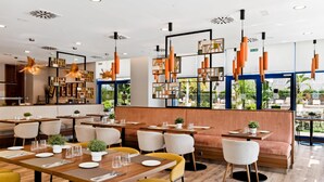 A modern restaurant interior with wooden tables, yellow and white chairs, and orange pendant lighting. - Hilton Garden Inn Sevilla (Seville)
