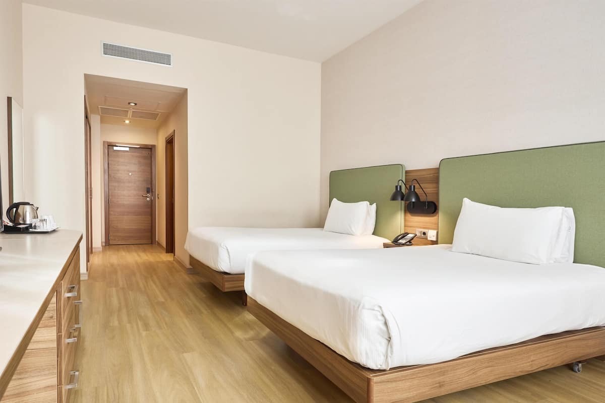 A hotel room with two beds, a wooden floor, a kitchenette, and a door leading to another room.