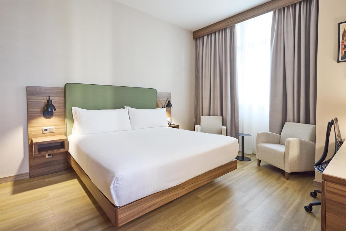 A hotel room with a large bed, a chair, a desk, a lamp, and a window with curtains.