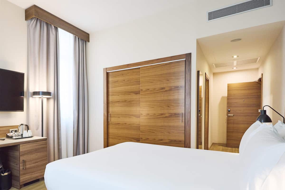 A hotel room with a large bed, wooden wardrobe, desk with a lamp, and a television.