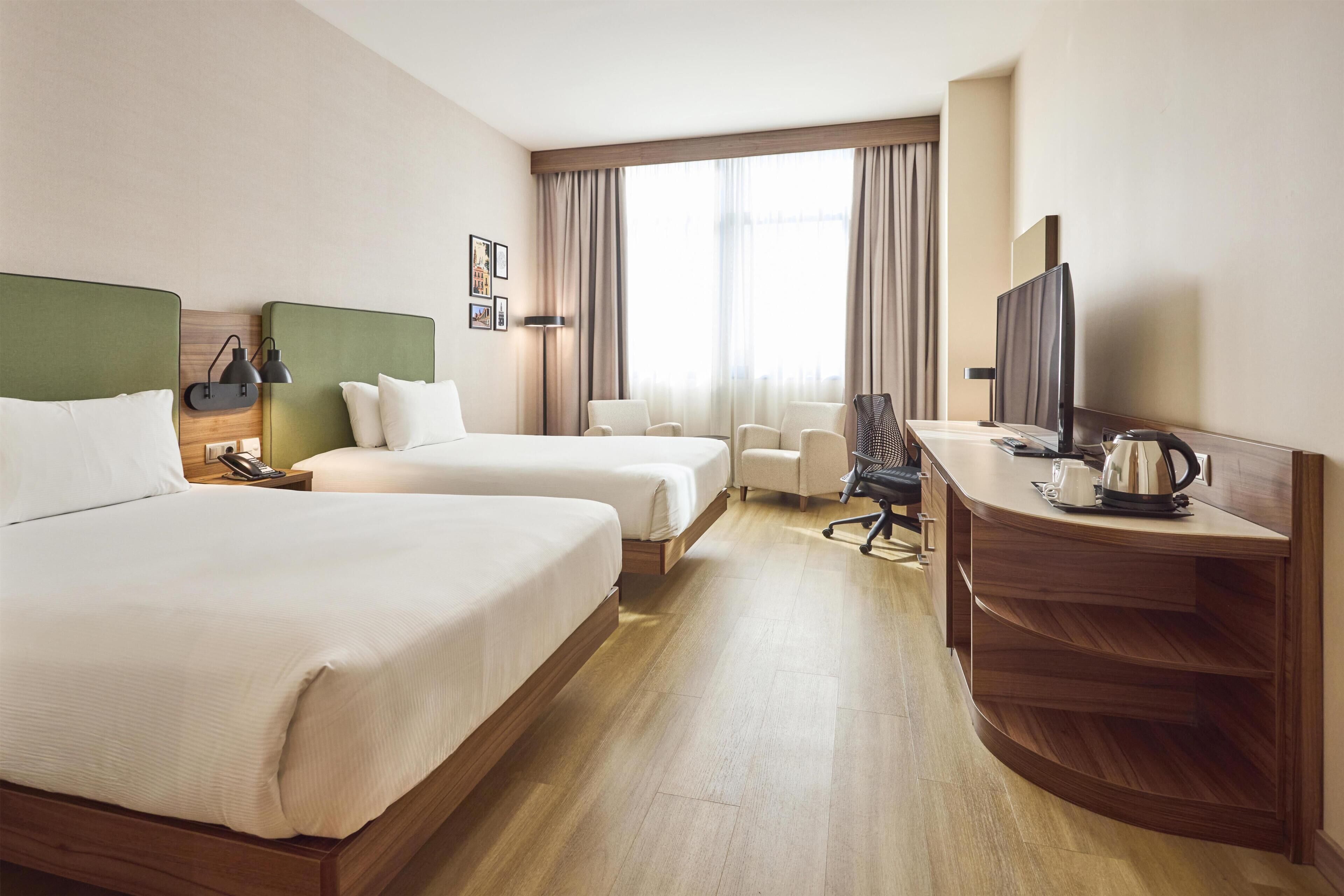 A hotel room with two beds, a desk with a television, a chair, and a window with curtains.