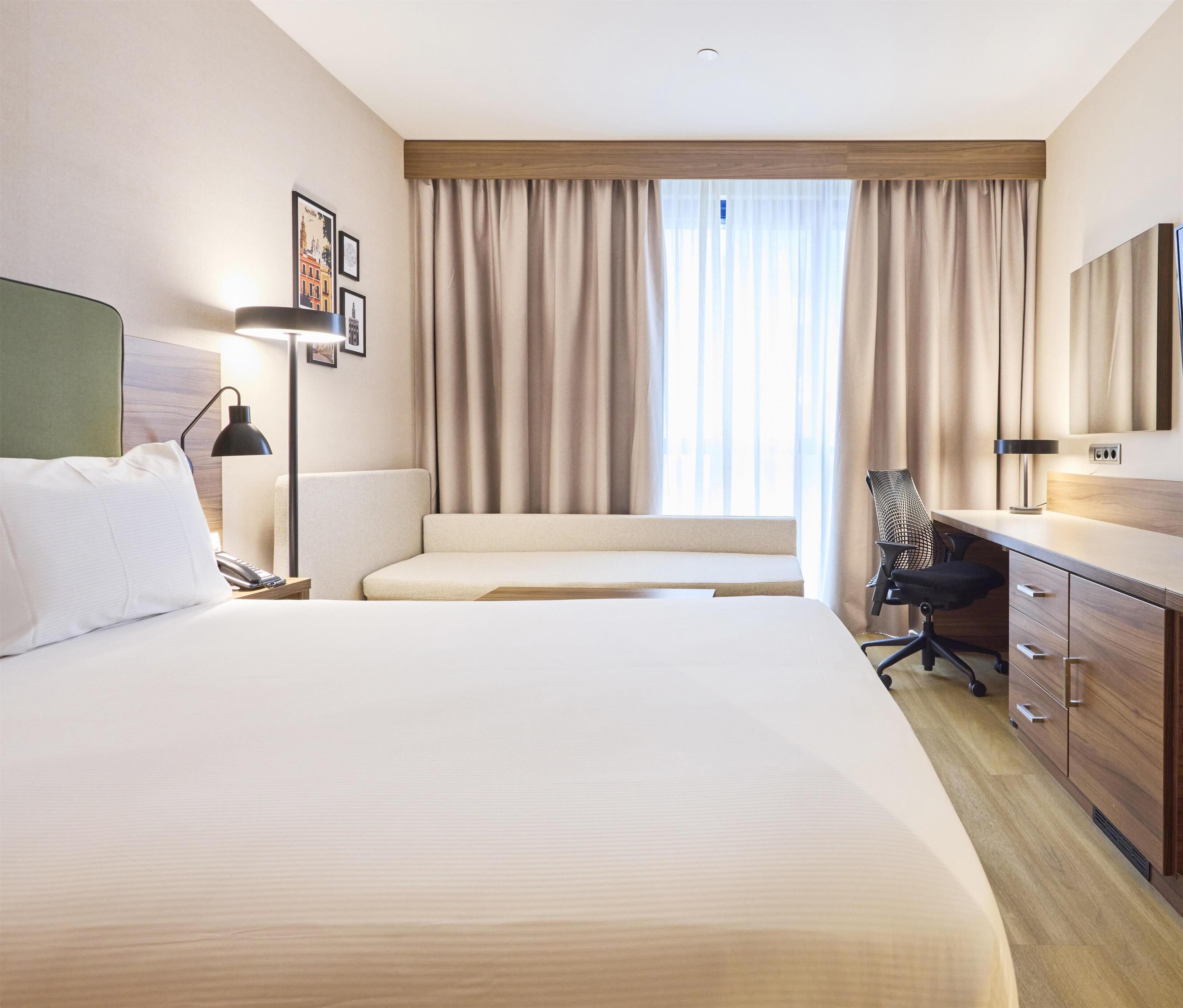 Een hotelkamer met een groot bed, een bureau, een stoel, een lamp en een raam met gordijnen.