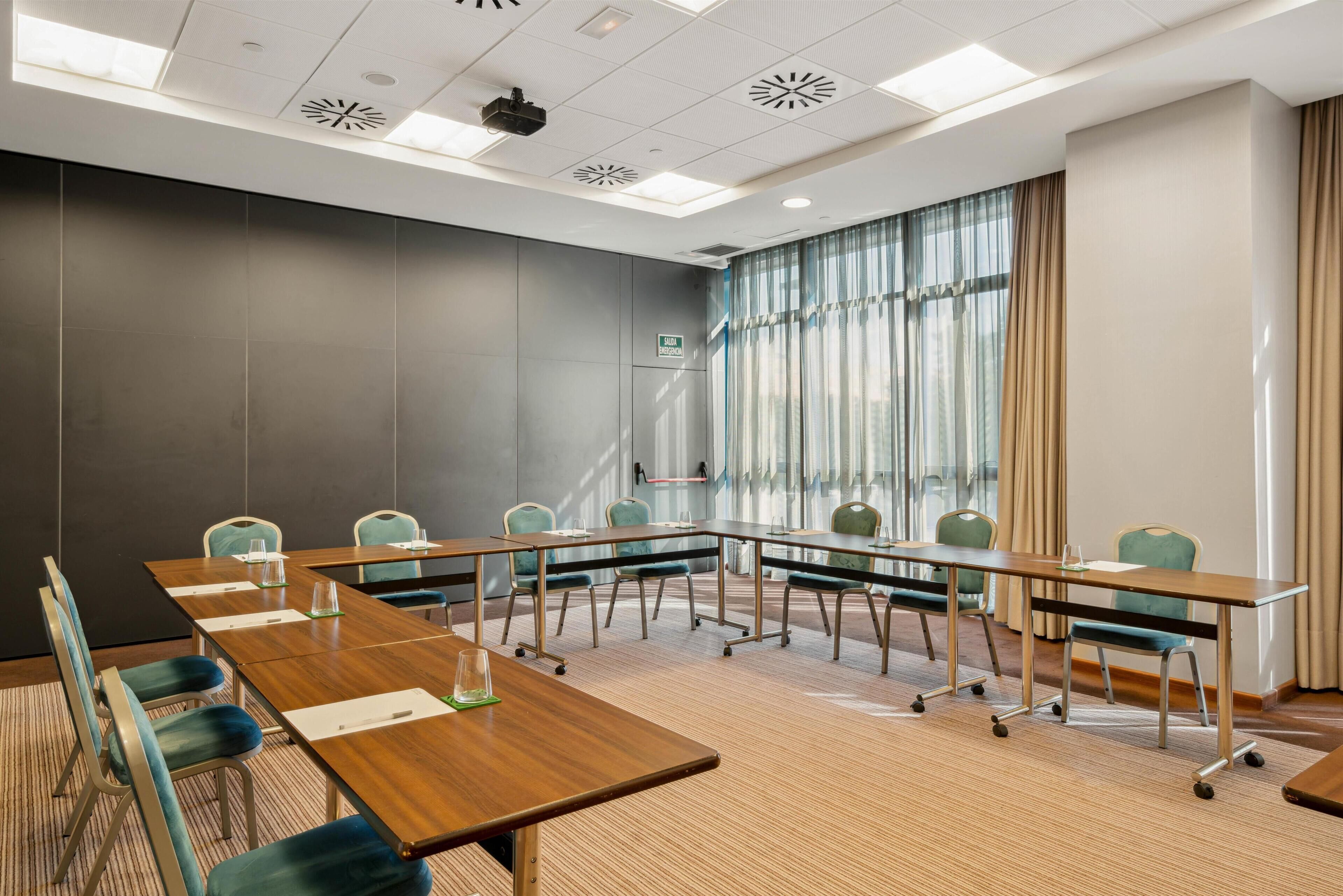 Une salle de conférence avec une longue table, des chaises, un projecteur et une fenêtre avec des rideaux.