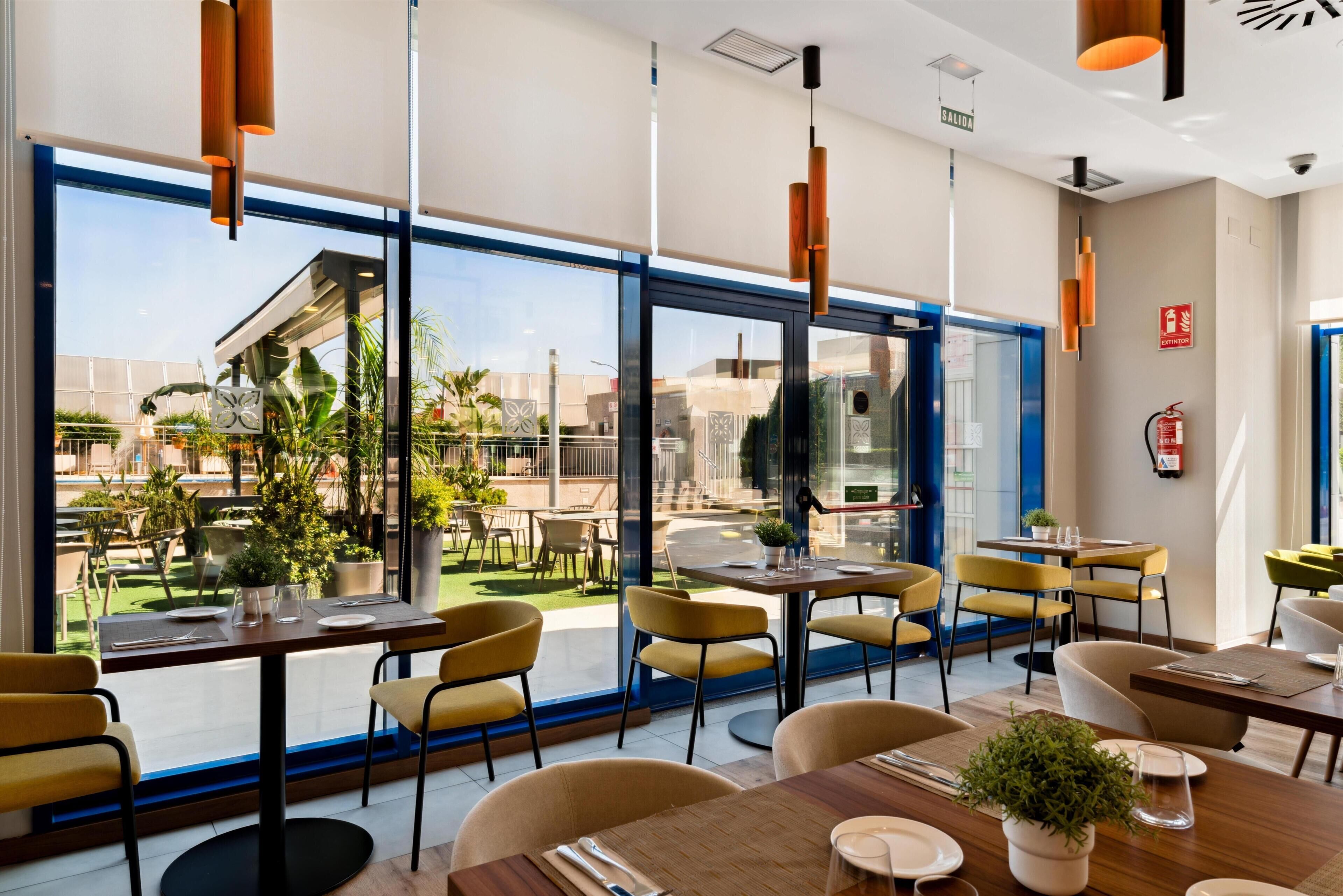 A modern restaurant with large windows, yellow chairs, and wooden tables.