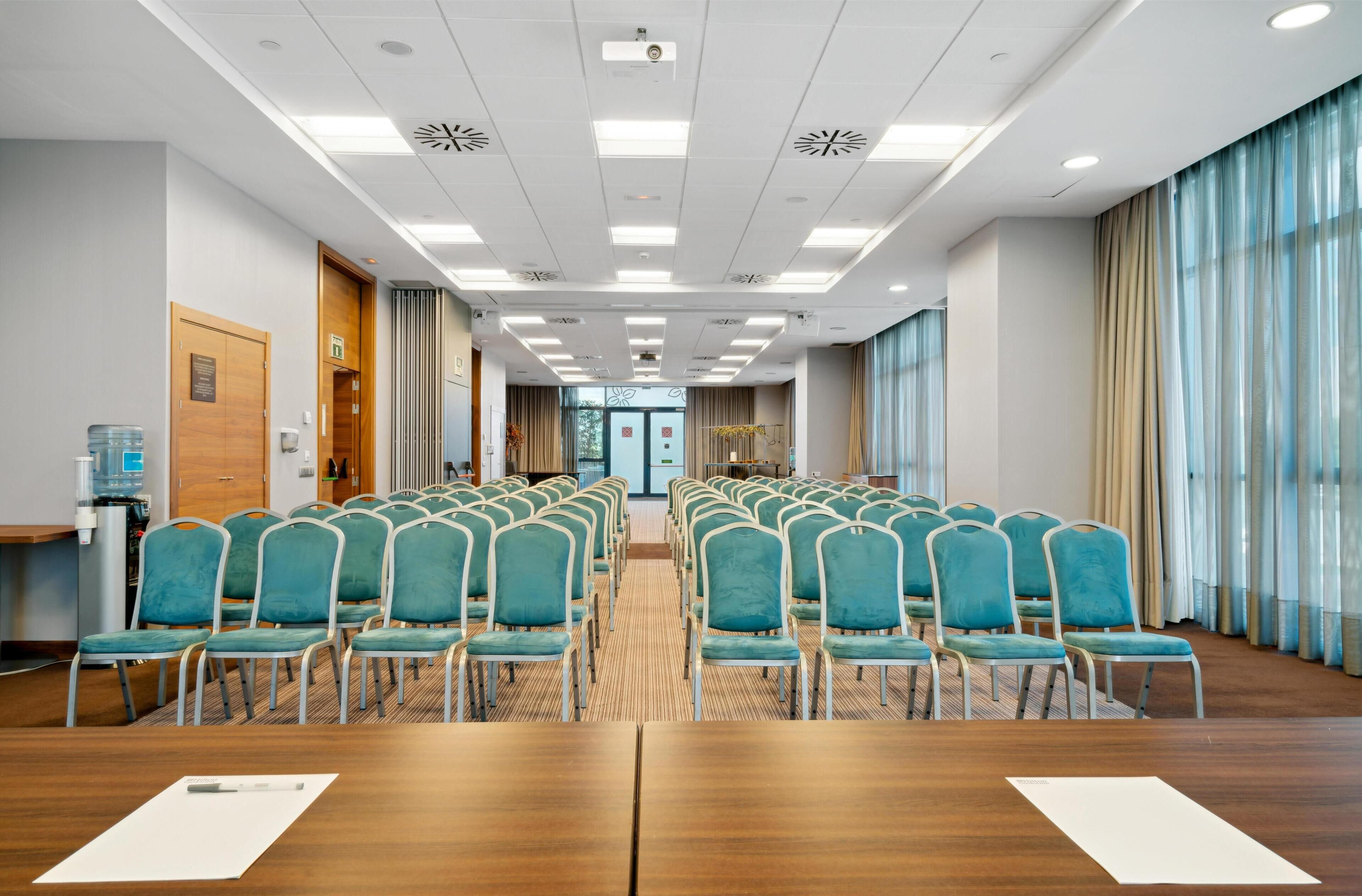 A conference room with rows of teal chairs, a wooden table, and a view through a glass door.