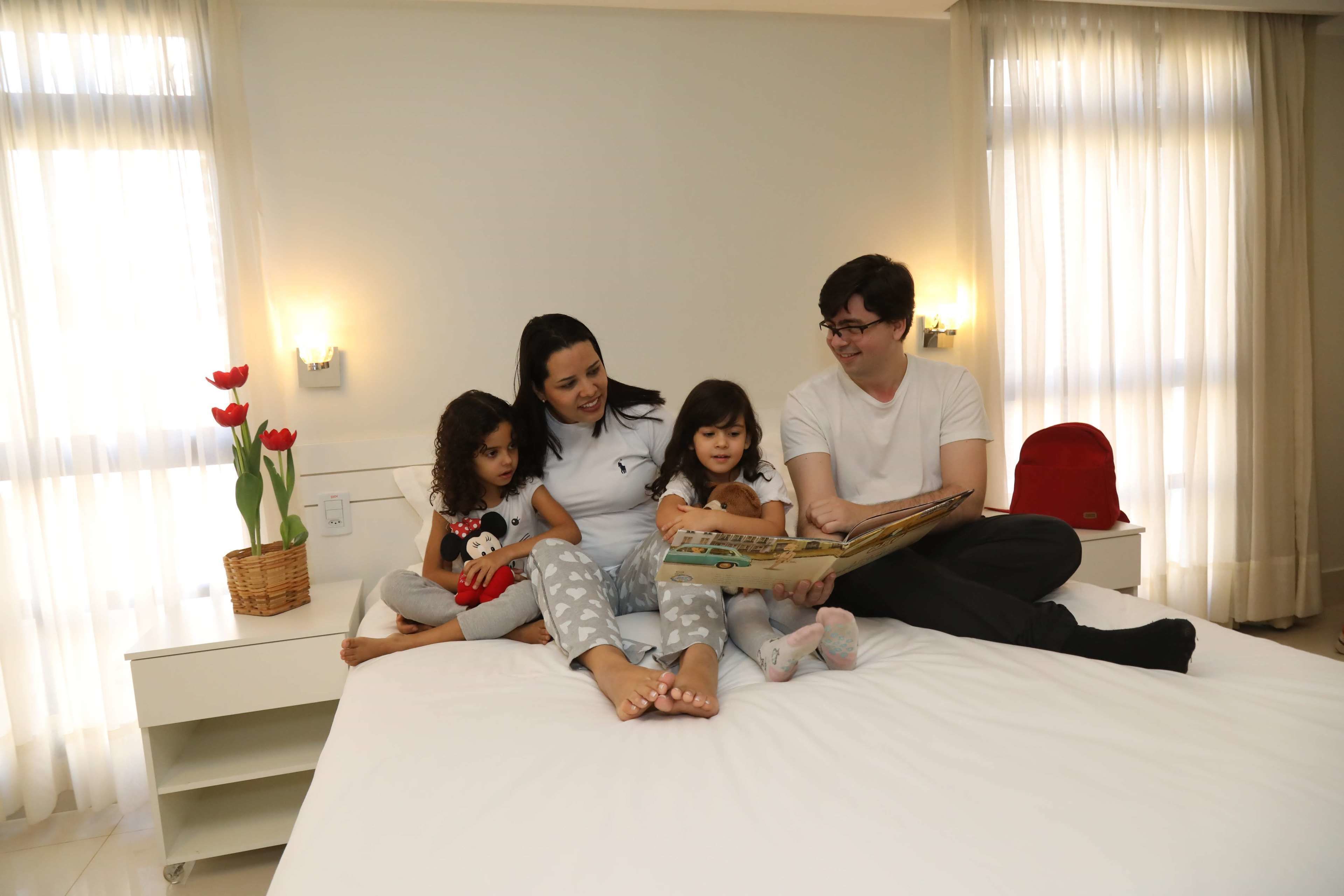 Eine Familie sitzt auf einem Bett in einem Zimmer mit einem weißen Nachttisch, einem Korb mit roten Blumen und einer Wandleuchte.