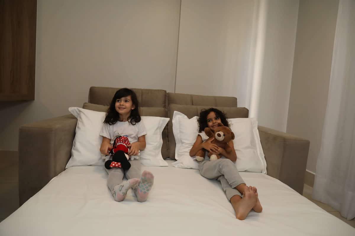 Two children sitting on a bed with pillows, holding stuffed animals.