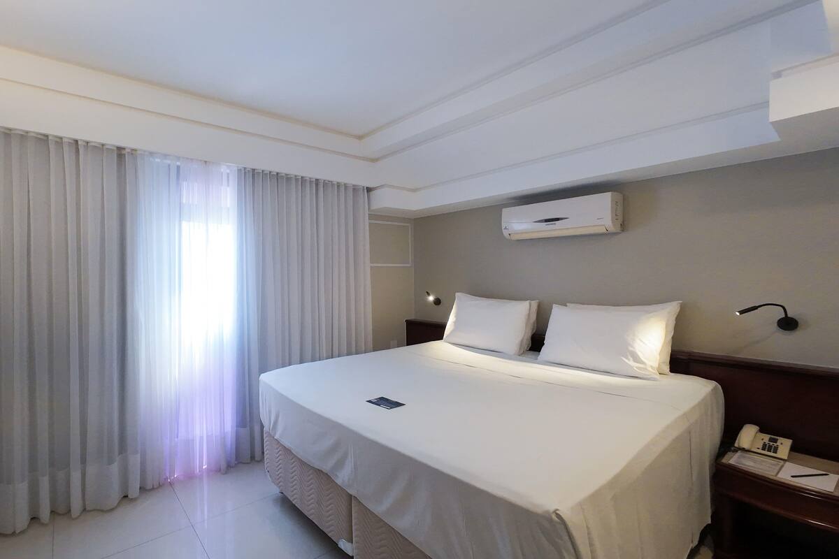 A hotel room with a bed, curtains, a wall-mounted air conditioner, a bedside table with a lamp, and a TV on the wall.