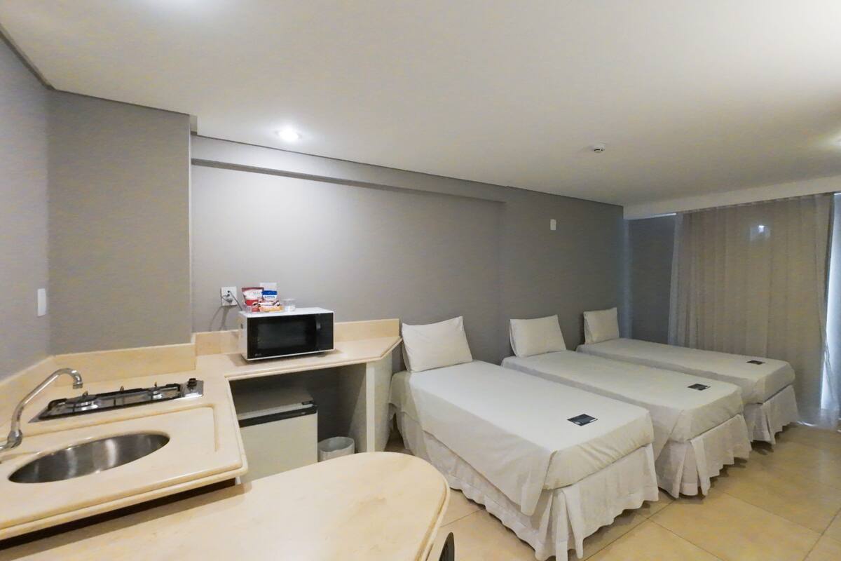 A hotel room with a kitchenette, two single beds, a TV, and a bathroom with a sink and toilet.