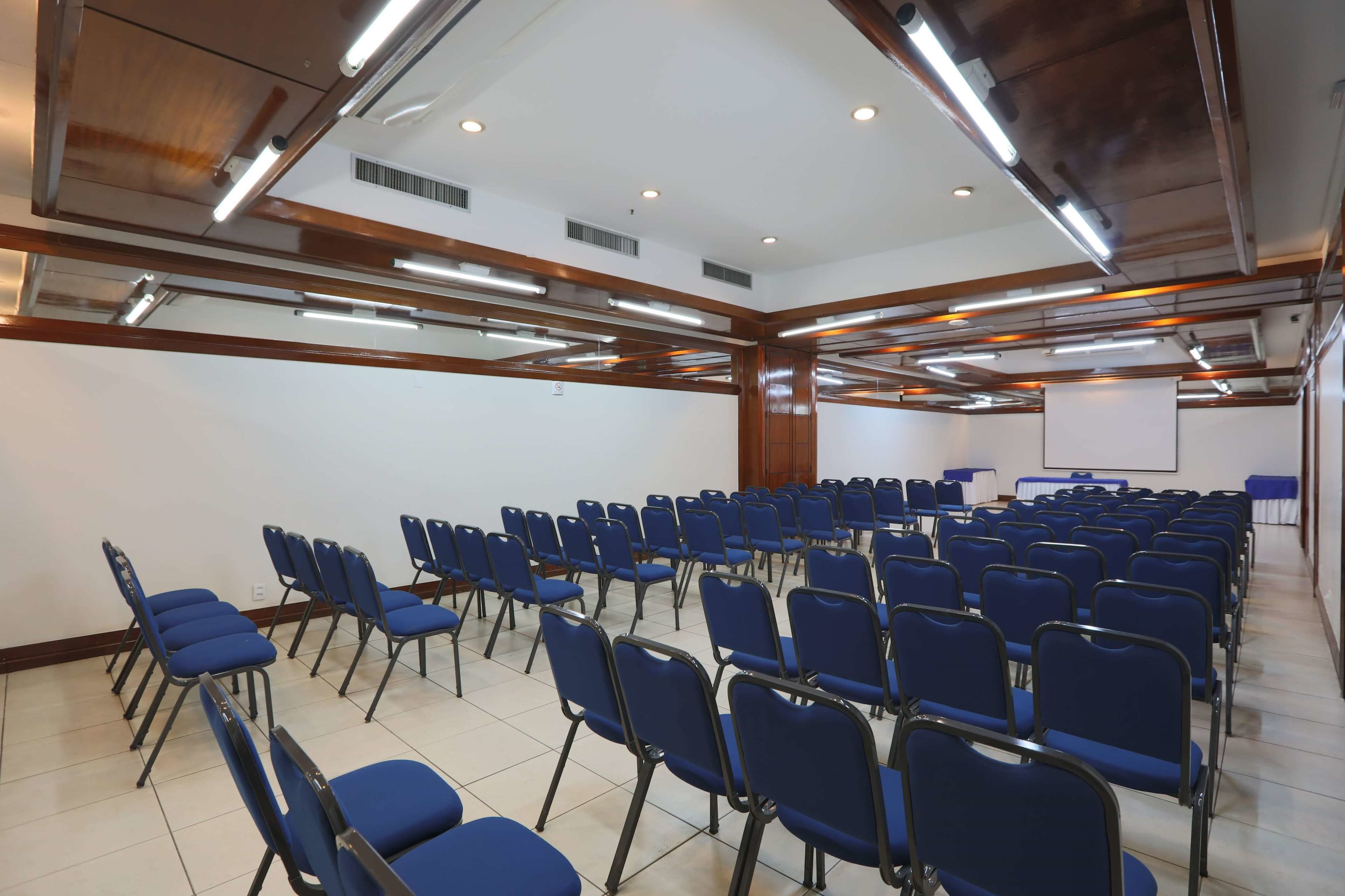 Uma sala de conferência com fileiras de cadeiras azuis, uma tela de projetor e uma lousa.