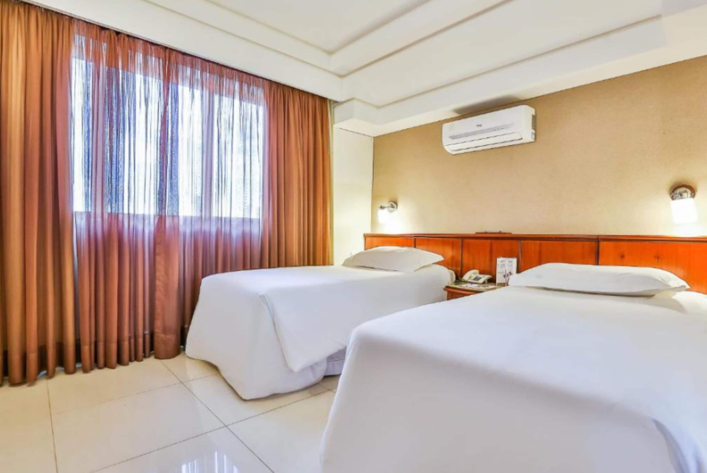 A hotel room with two beds, a window with curtains, an air conditioning unit, and a bedside table with a phone.