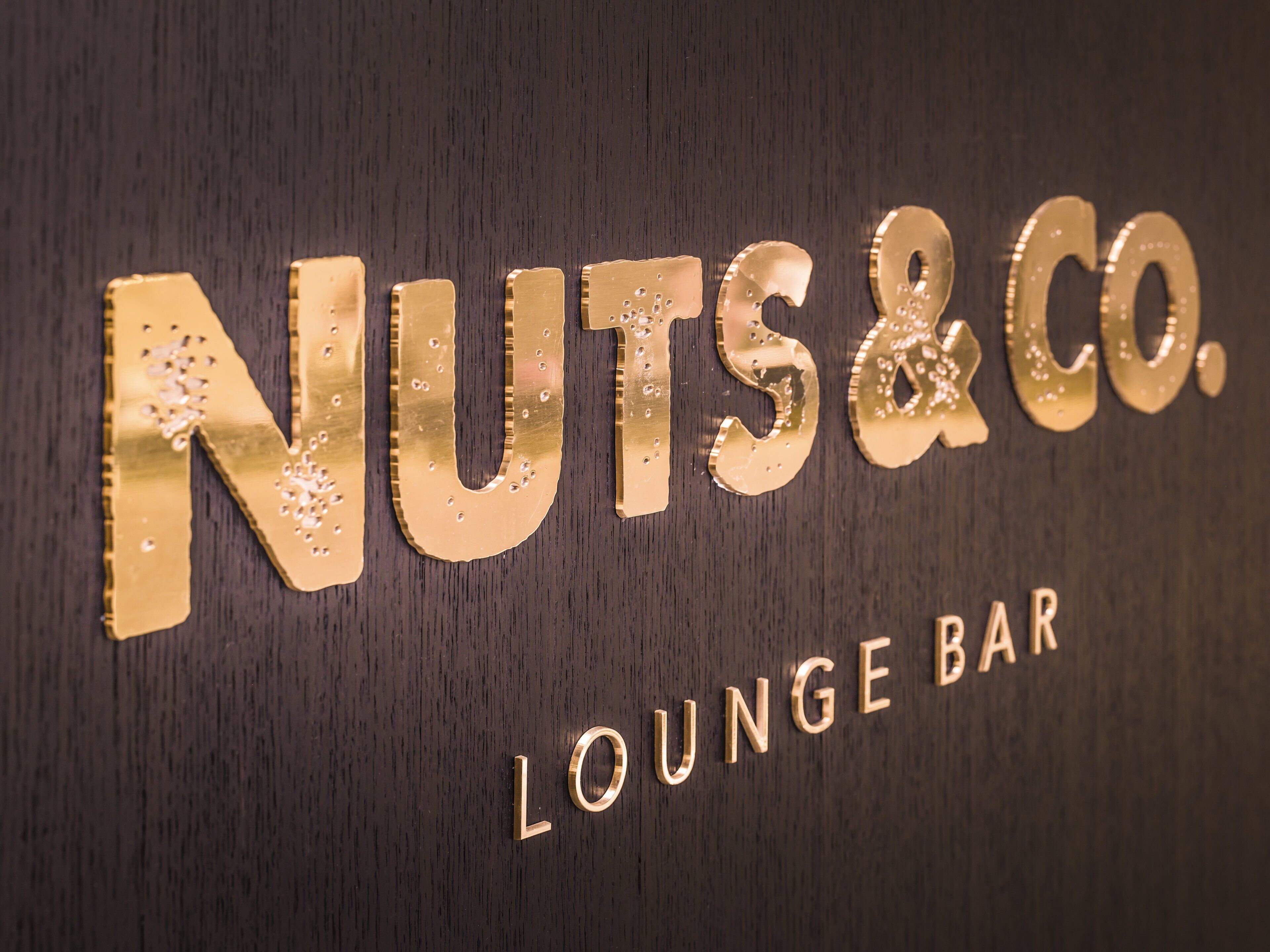 A sign with the text "NUTS&CO. LOUNGE BAR" on a dark wooden background.