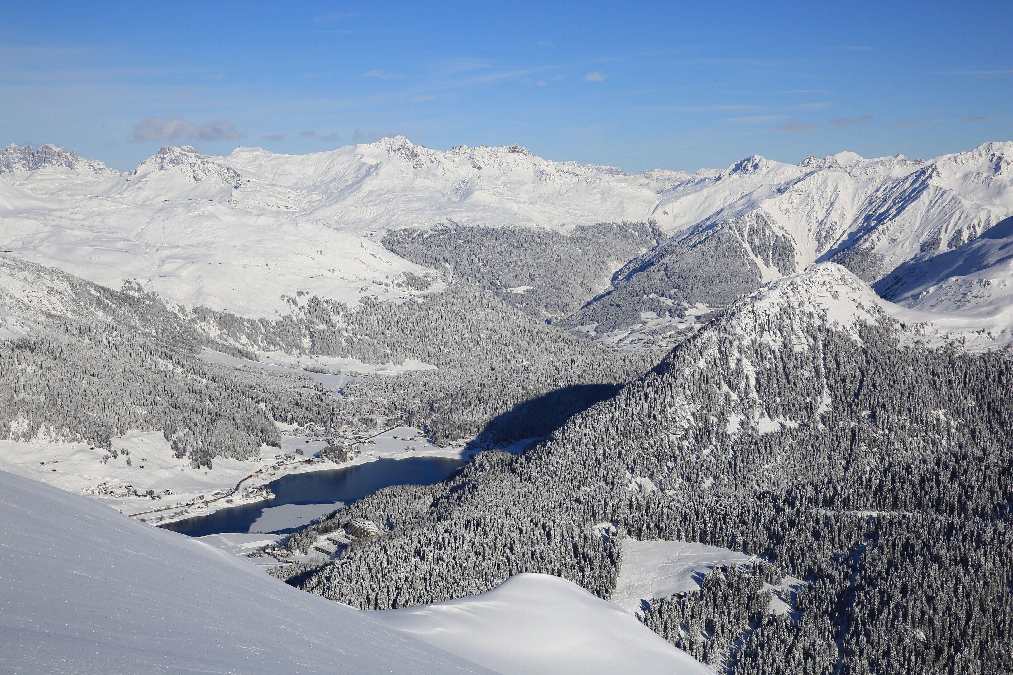 A panoramic view of a snow-covered mountain range with a valley and a lake.