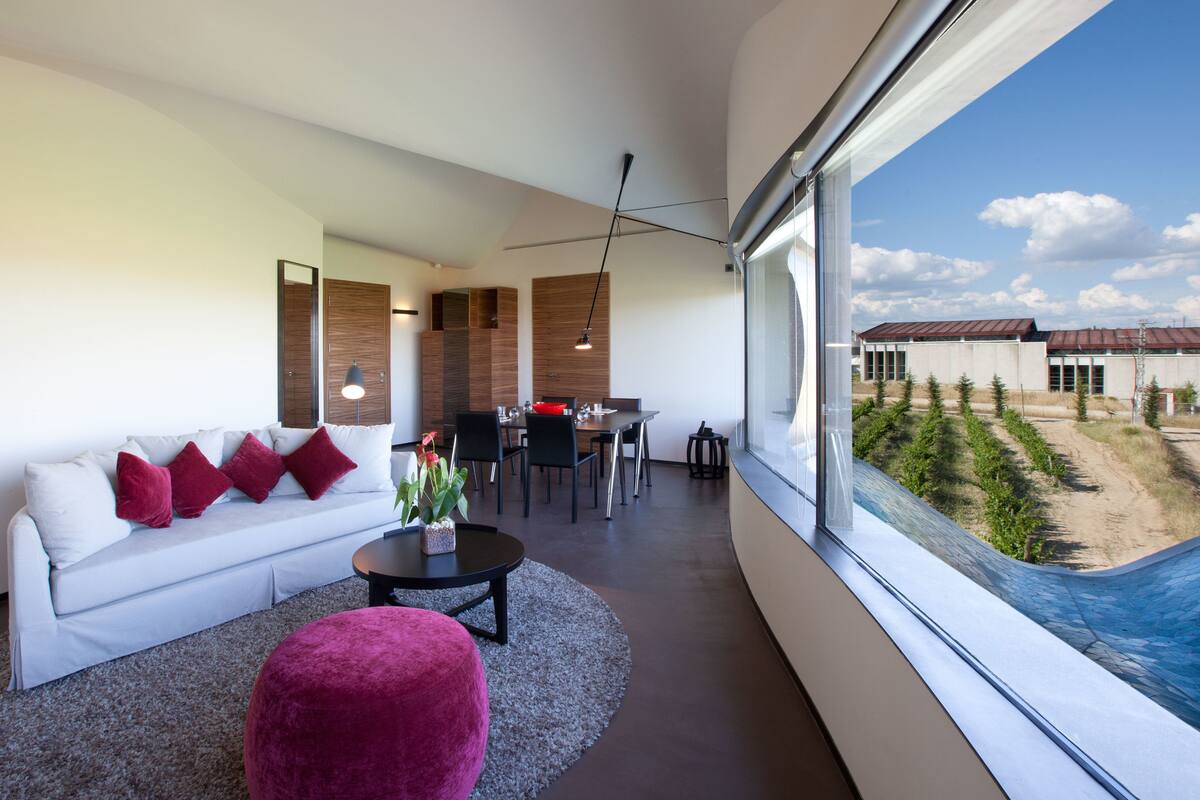 Un salon moderne avec un canapé blanc, des coussins rouges, une table basse ronde et une vue sur un immeuble et la verdure environnante.