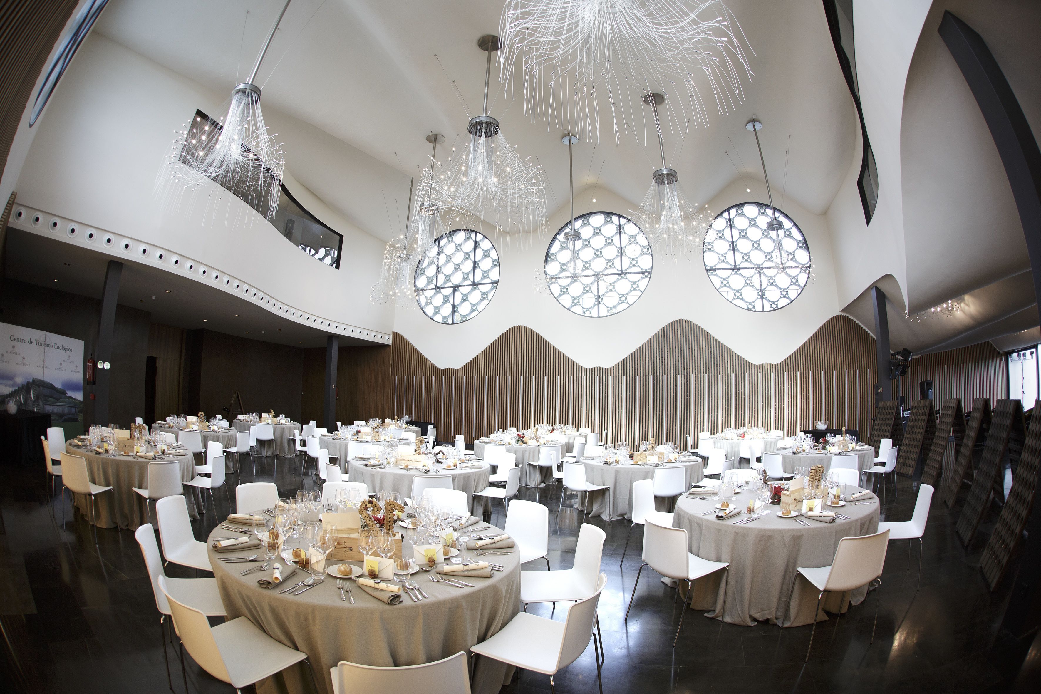 A banquet hall with round tables set for dining, large windows, and modern chandeliers.