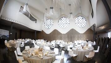 Un salón de banquetes con mesas redondas dispuestas para comer, ventanas amplias y candelabros modernos.