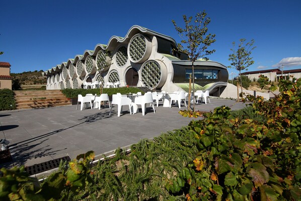 A modern building with unique circular windows and a curved roof, surrounded by white chairs and greenery.