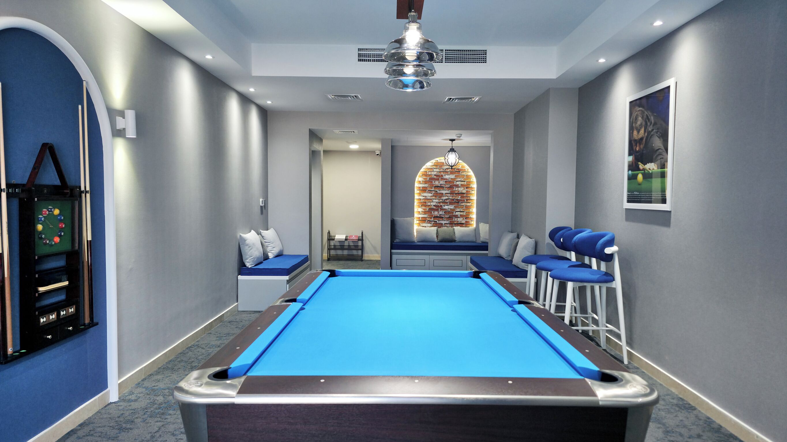A pool table with a blue felt surface, a seating area with blue chairs, a wall clock, and a ceiling fan.