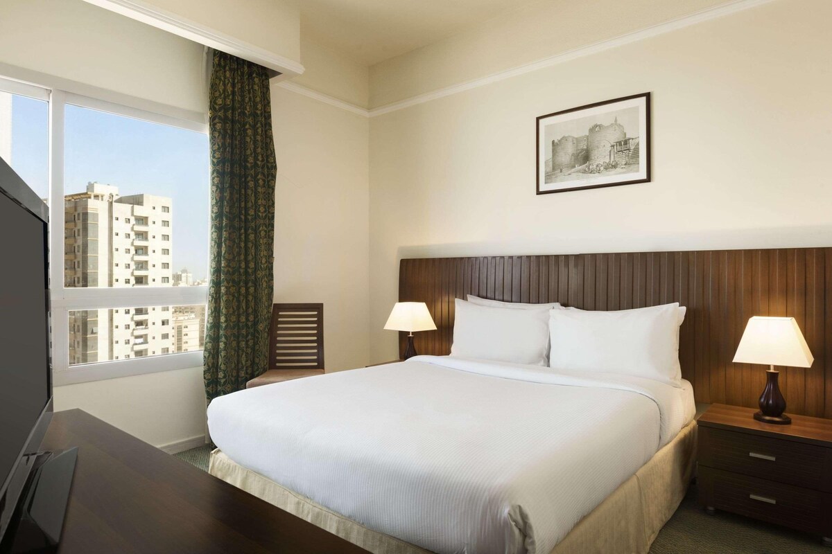 A hotel room with a large bed, two bedside lamps, a framed picture on the wall, and a view of buildings through the window.