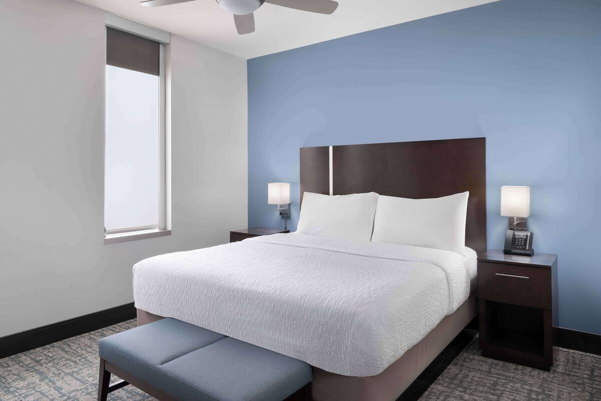 Een hotelkamer met een groot bed, twee nachtkastjes met lampen, een bankje en een plafondventilator.