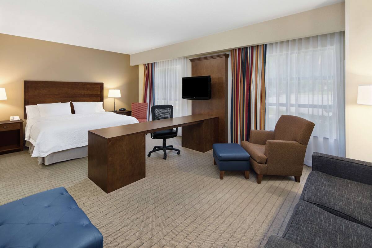 A hotel room with a bed, desk, two chairs, a TV, and a large window with curtains.