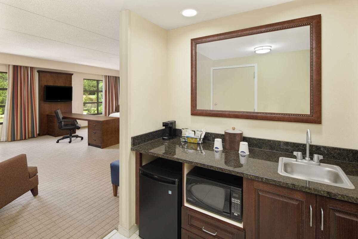 A hotel room with a kitchenette, a large mirror, a flat-screen TV, a desk with a chair, and a view of the outdoors.