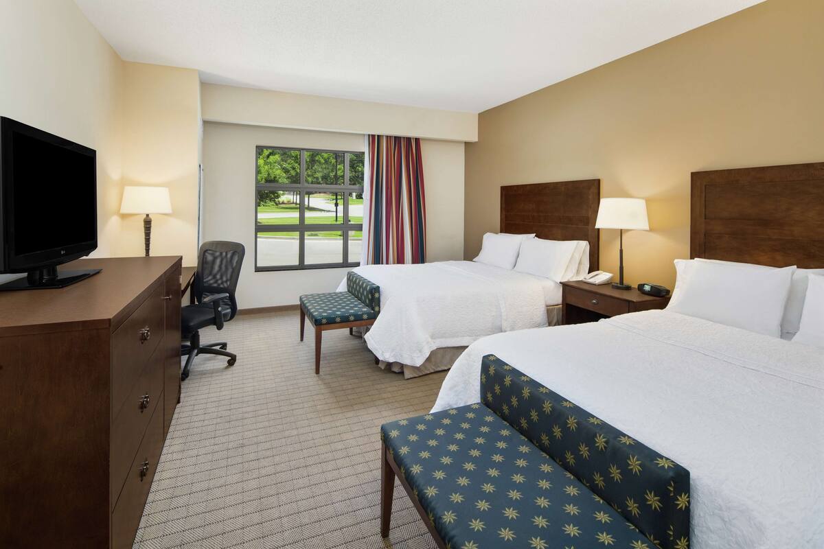 A hotel room with two beds, a desk with a TV, a chair, a lamp, and a window with a view of greenery.
