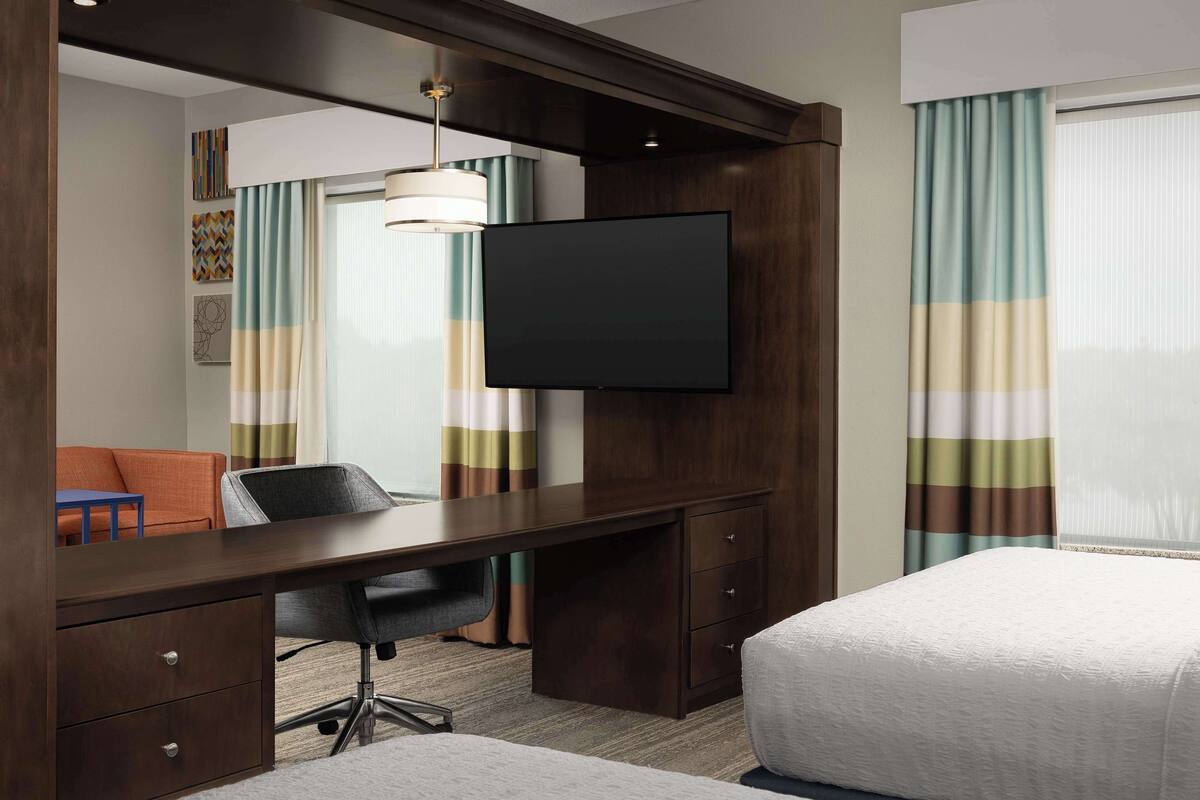 A hotel room with a wooden desk, a flat-screen TV, a bed, and a window with curtains.