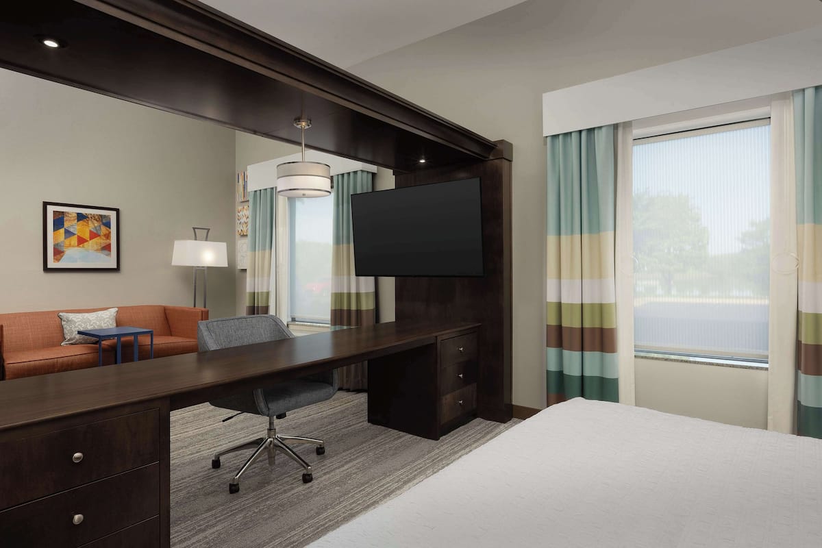 A hotel room with a bed, a desk with a chair, a TV, a sofa, and a window with curtains.