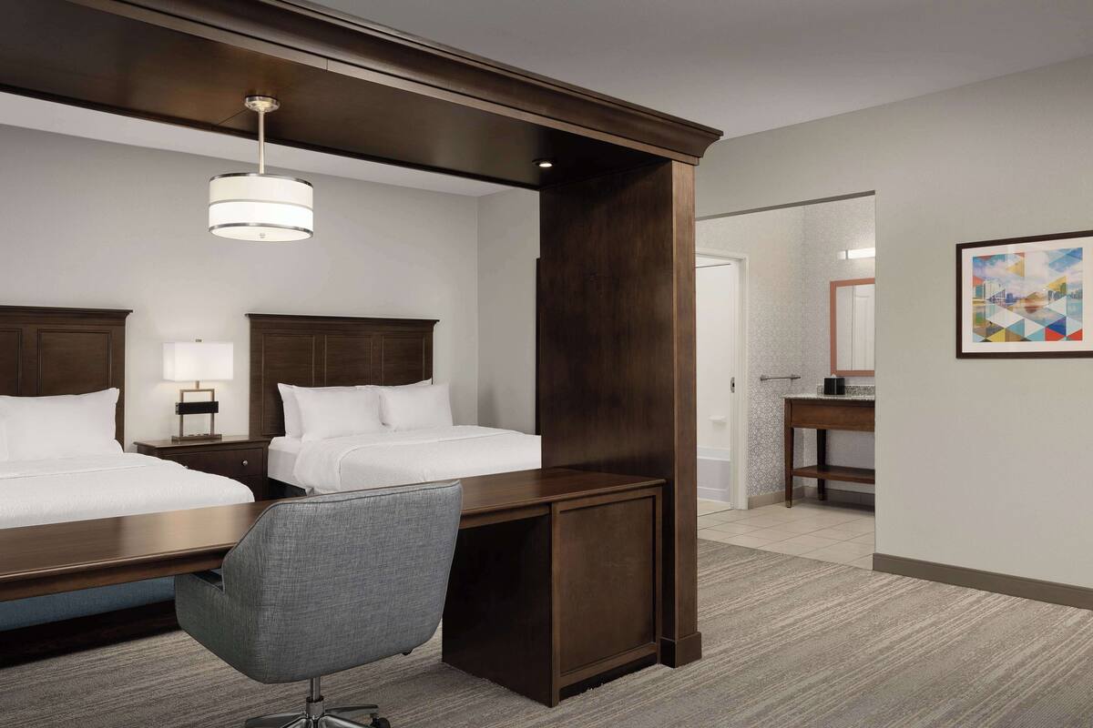 A hotel room with two beds, a desk, a chair, a lamp, and a painting on the wall.