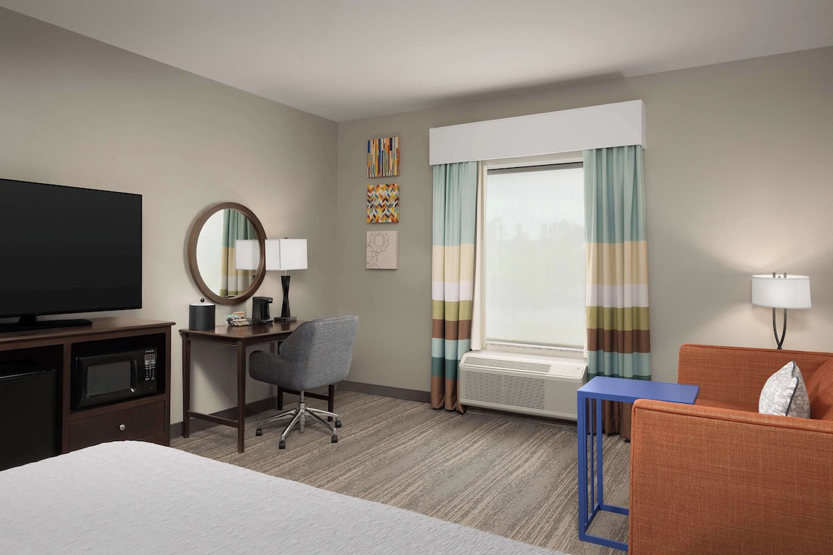 A hotel room with a bed, a desk with a chair, a television, a microwave, a small table, and a window with curtains.