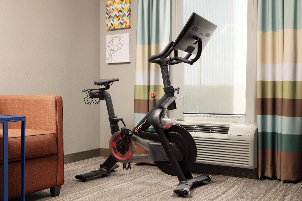 A Peloton exercise bike in a room with a blue chair, a radiator, and a window with curtains.