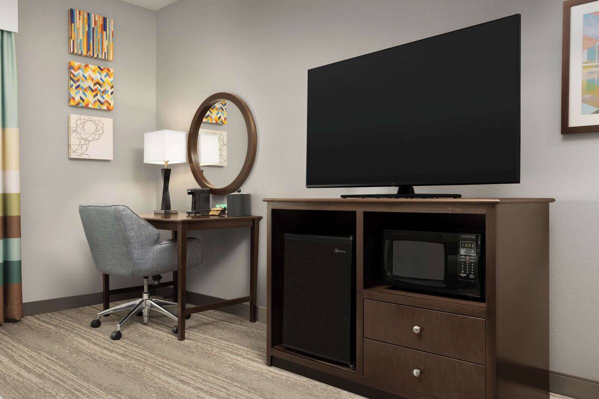 A hotel room with a flat-screen TV, a desk with a lamp, a microwave, and a refrigerator.