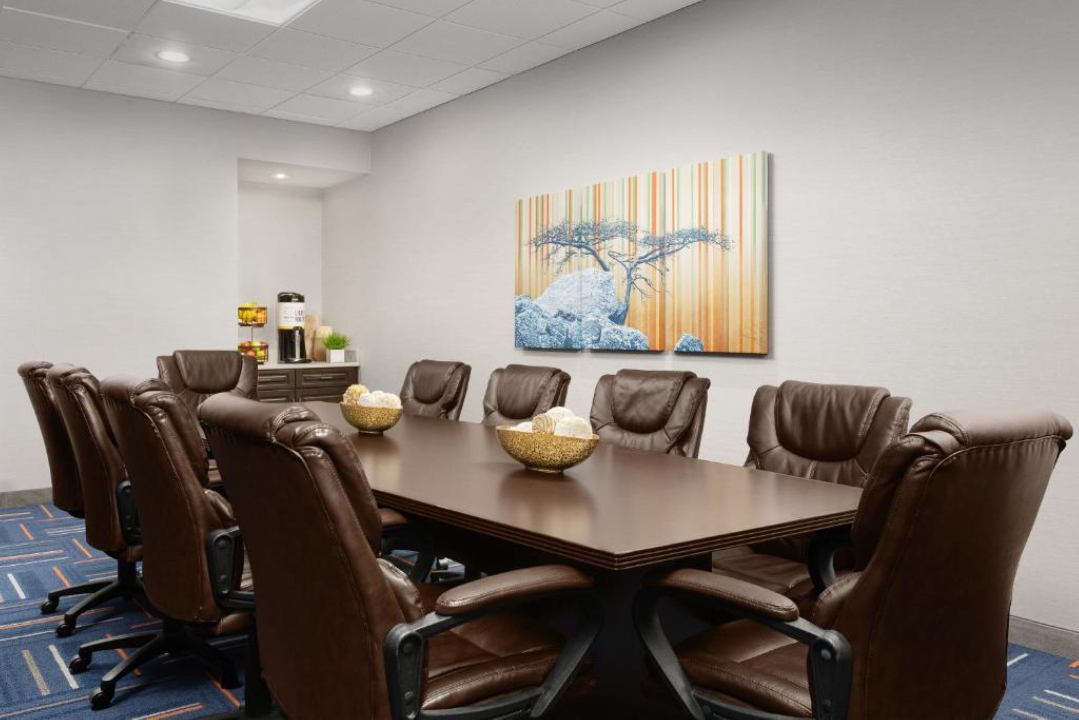 A conference room with a long table, eight brown leather chairs, and a large painting of a tree on the wall.