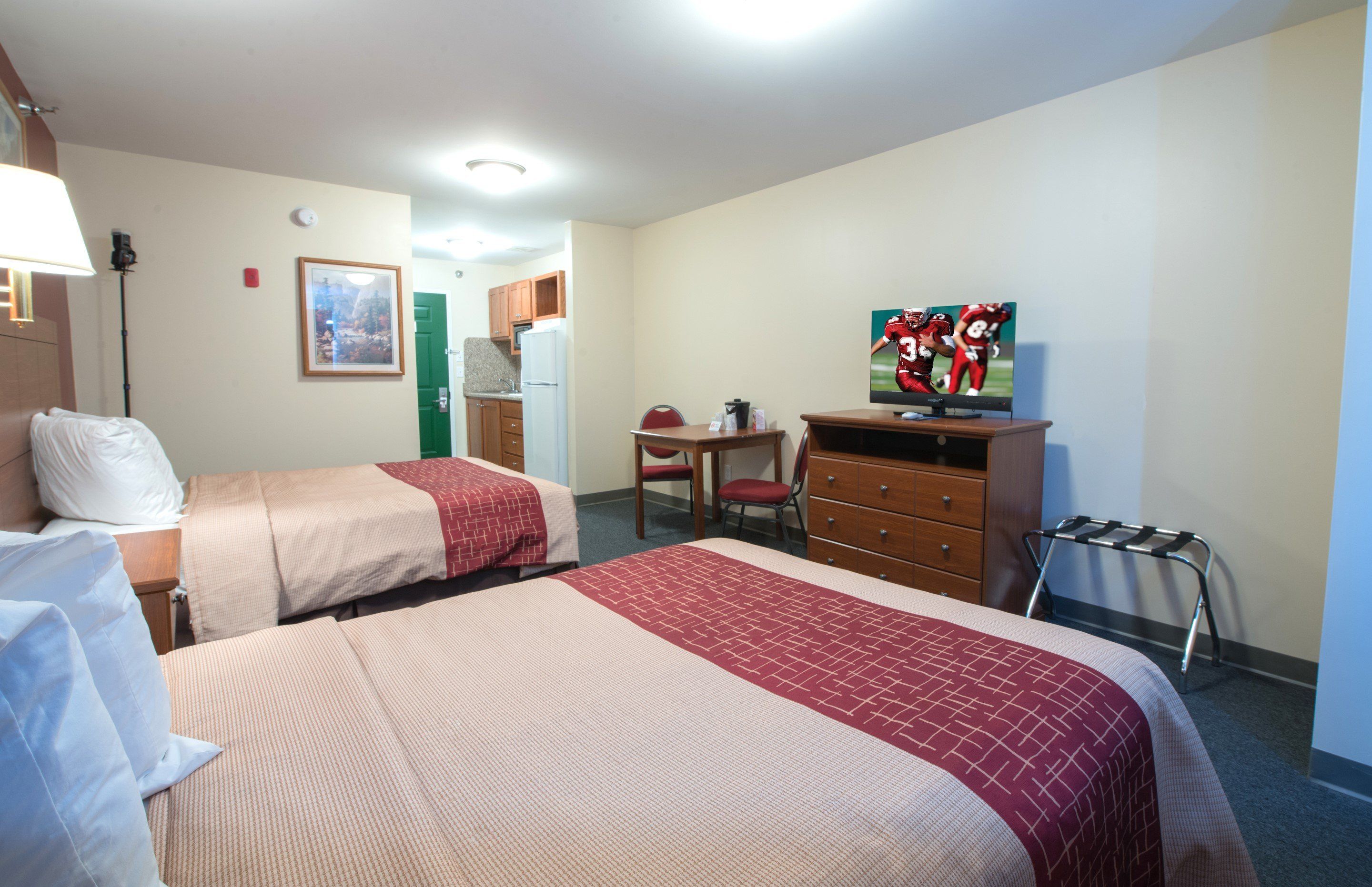 A hotel room with two beds, a desk, a television, and a kitchenette.