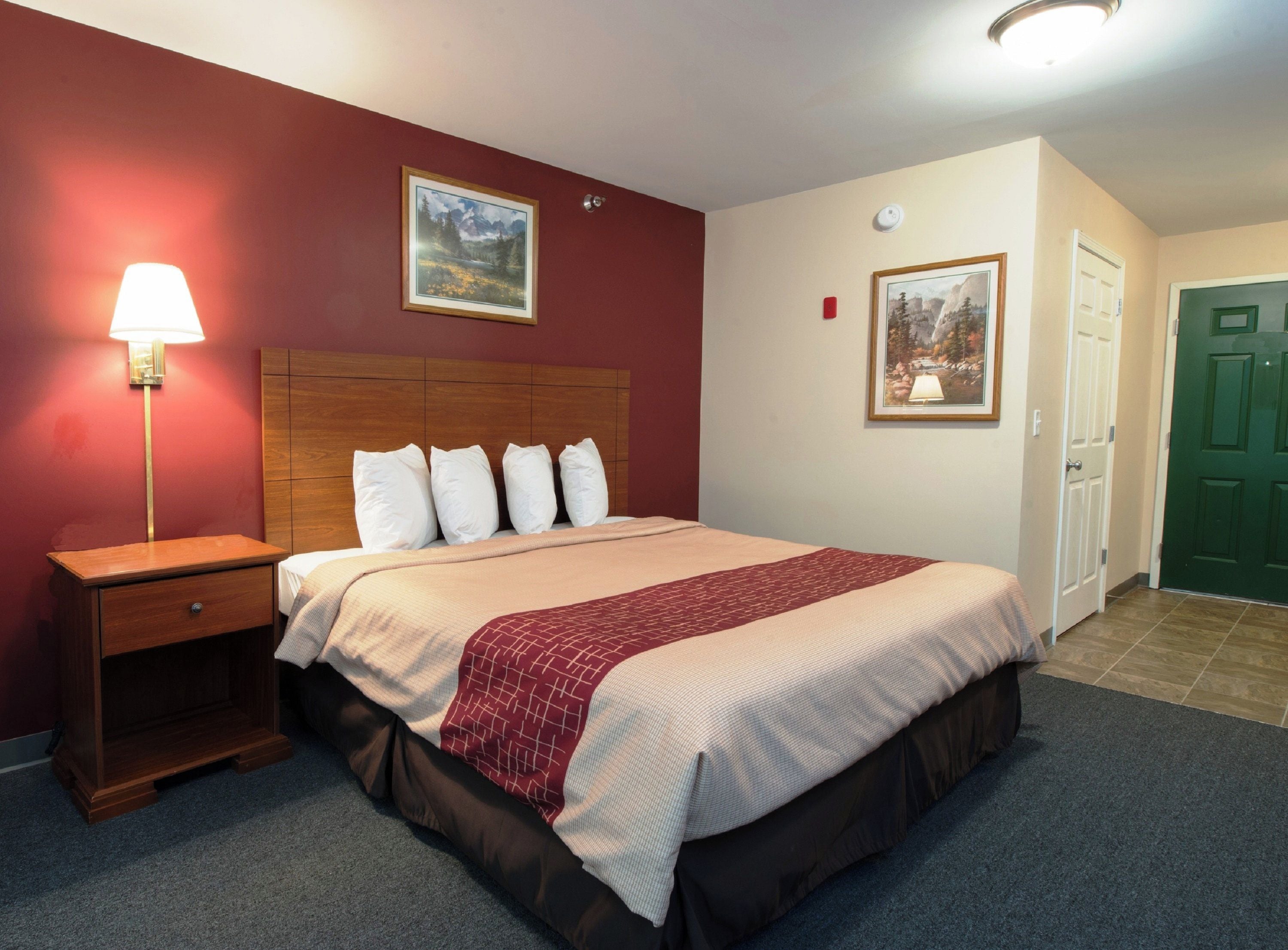 A hotel room with a bed, a nightstand, a lamp, two paintings on the wall, and a green door.