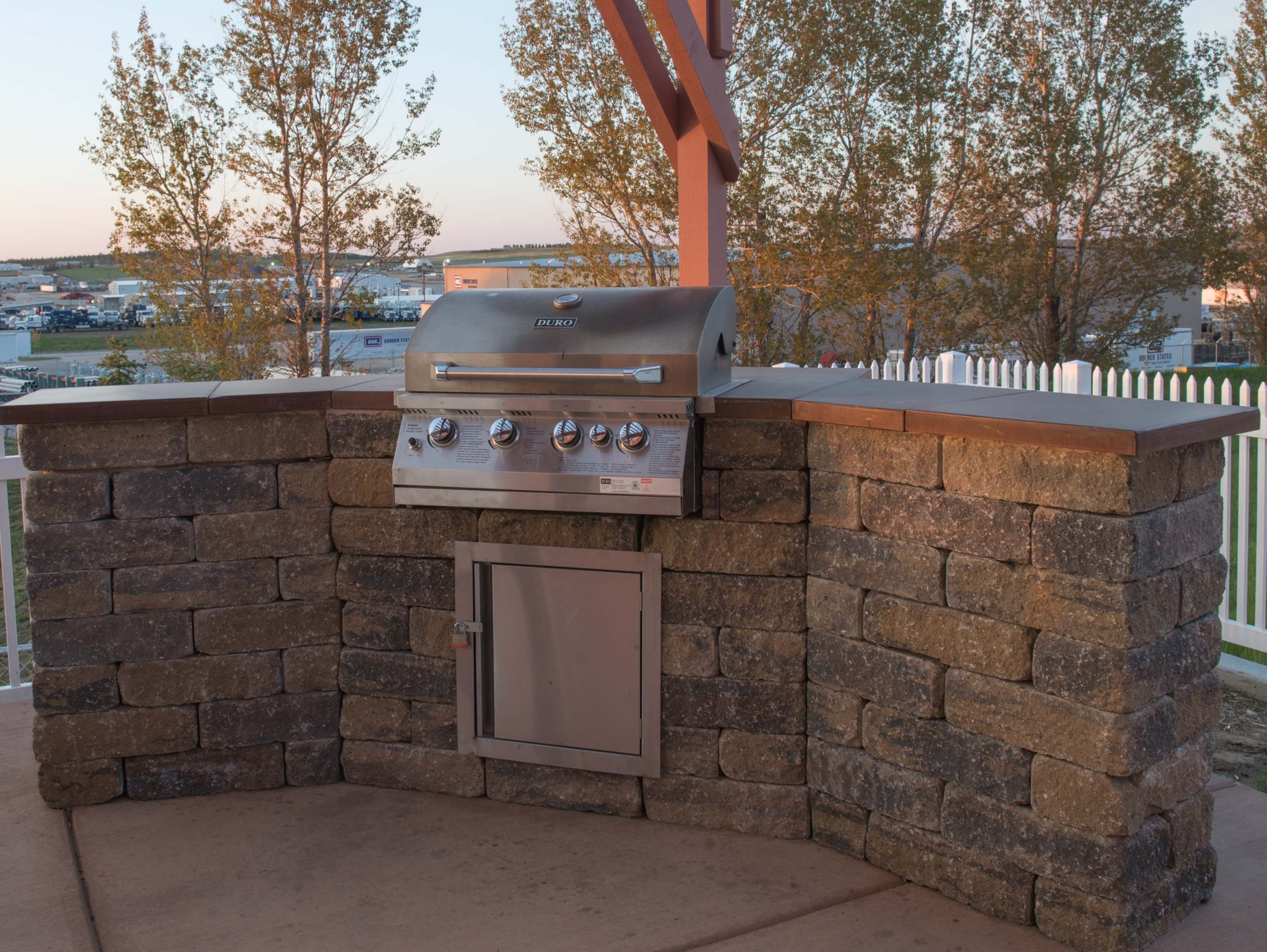 Un barbecue extérieur, avec un mur en pierre et une porte en métal, se dressant sur un fond de verdure et offrant une vue lointaine sur des bâtiments.
