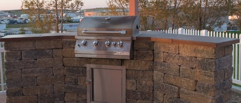 An outdoor grill with a stone wall and a metal door, set against a backdrop of trees and a distant view of buildings.
