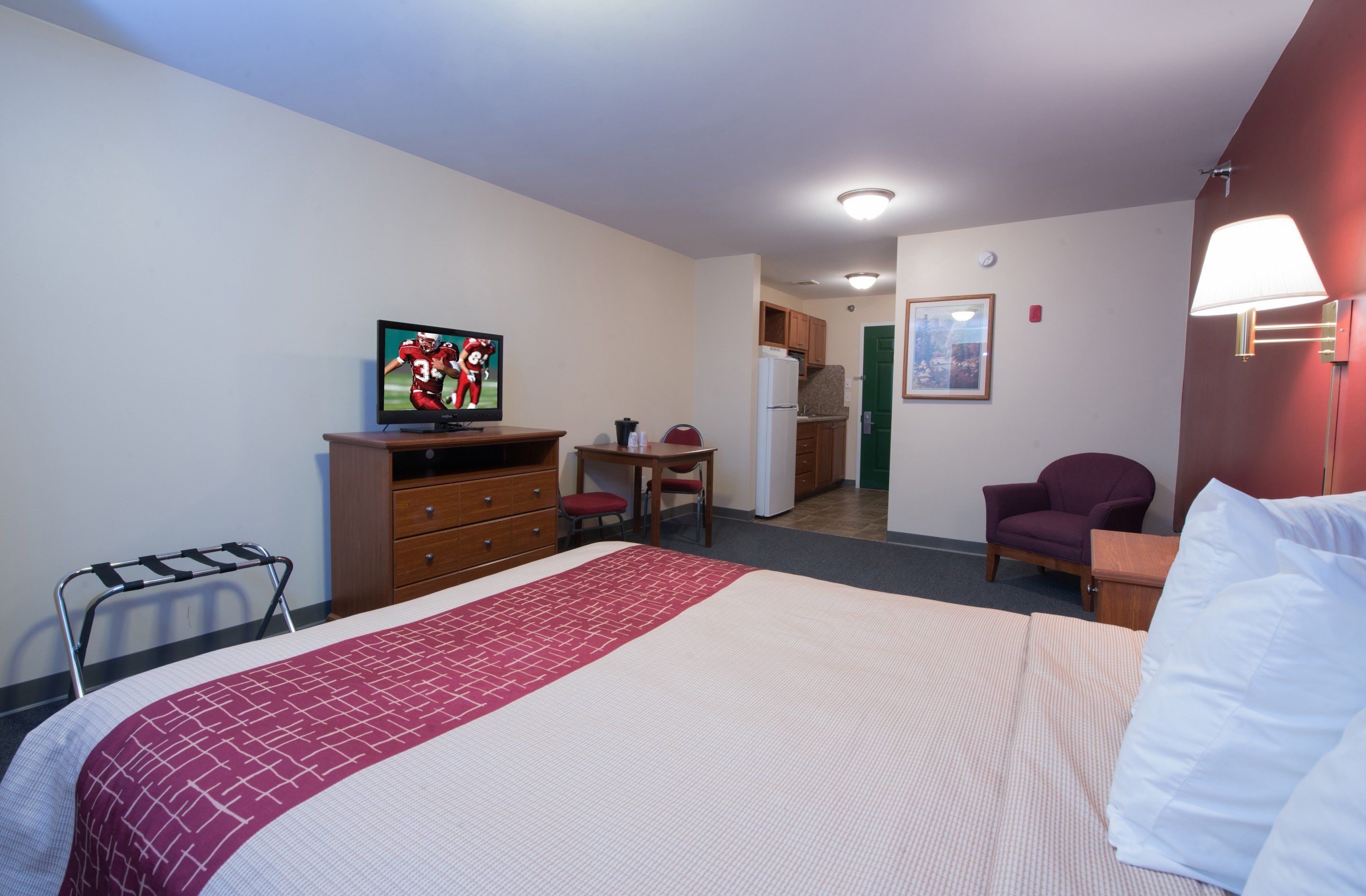 A hotel room with a bed, a desk, a chair, a television, a refrigerator, and a door leading to a kitchen.