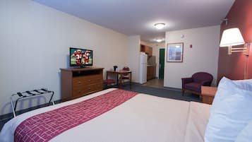 A hotel room with a bed, a desk, a chair, a television, a refrigerator, and a door leading to a kitchen.
