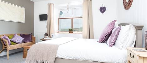 A bedroom with a bed, a bedside table, a chair, a window, and a wall-mounted TV.